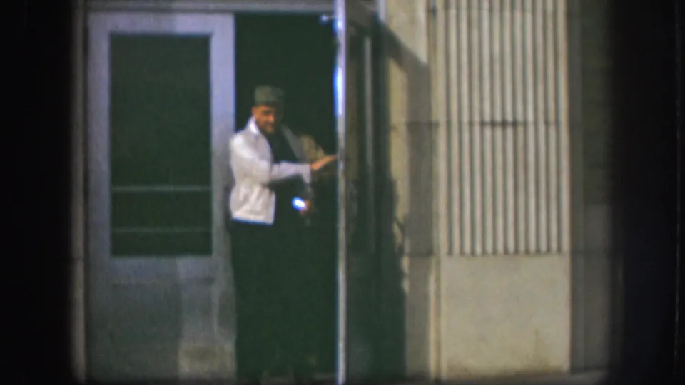 A man in a white jacket and dark cap exits through a green door on a corrugated metal commercial building, Kansas, circa 1958, 8mm.