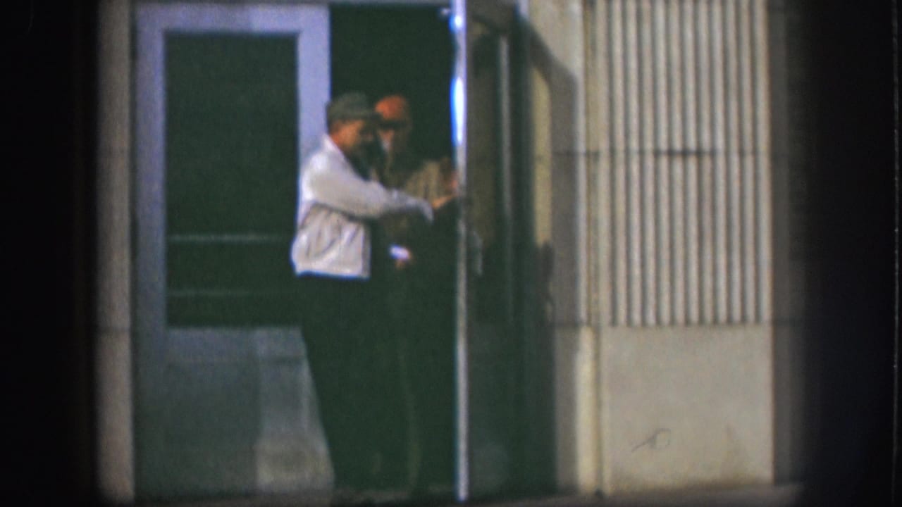 Two men at a building doorway; the man in a light shirt appears to face or address the other man in a dark jacket.