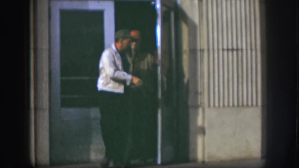Two men emerge from a building entrance, Kansas, circa 1958; one in a light shirt looks downward while the other in a dark jacket stands beside him.