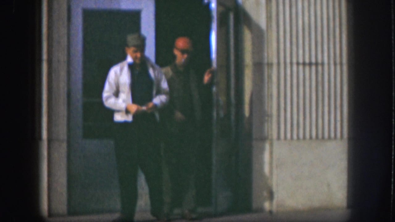 Two men pause near a glass-panel door and ribbed-siding building exterior, consistent with Kansas 1958 per source metadata; one wears a brimmed hat and tie.