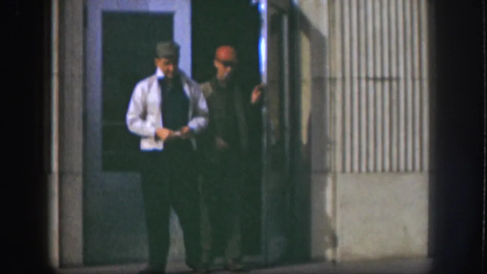 Two men pause near a glass-panel door and ribbed-siding building exterior, consistent with Kansas 1958 per source metadata; one wears a brimmed hat and tie.