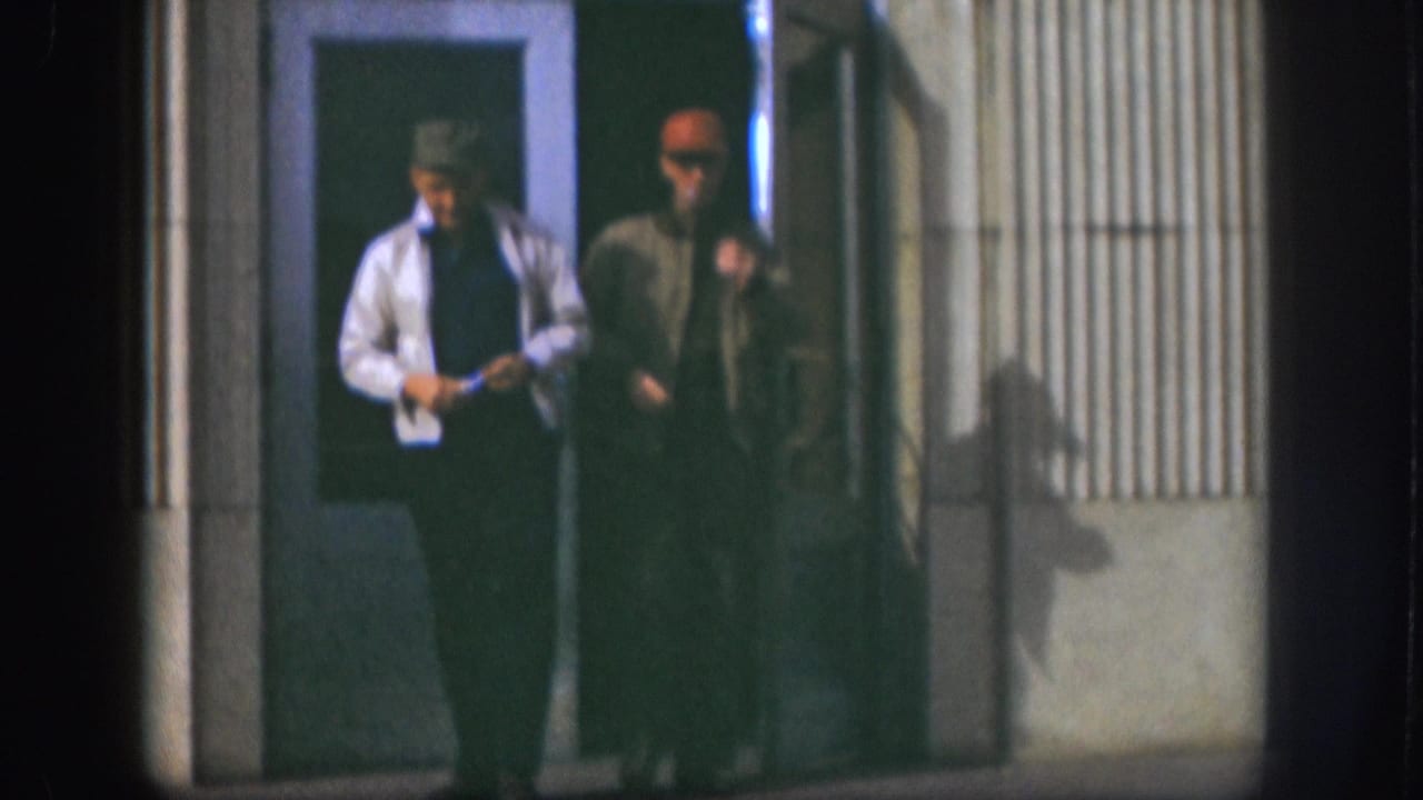 Two men near a building door; the one in a white shirt and brimmed hat looks down, the other in a red cap stands beside him.