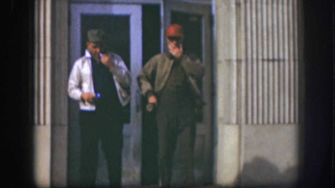 Two middle-aged men in baseball caps and work jackets stand at a glass-door building entrance smoking cigarettes; Kansas, circa 1958, 8mm color film.