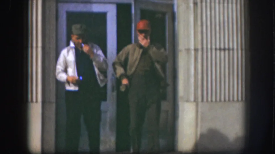 Two middle-aged men in baseball caps and work jackets stand at a glass-door building entrance smoking cigarettes; Kansas, circa 1958, 8mm color film.