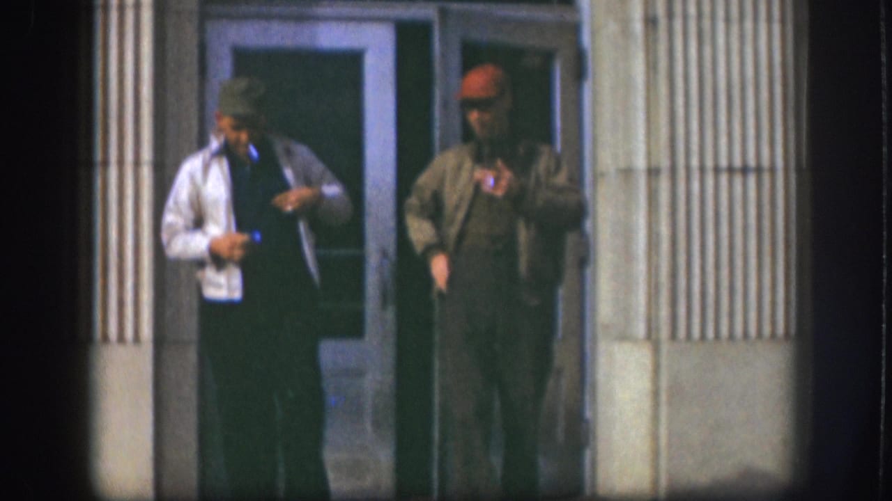 Two middle-aged men stand outside a building with fluted pilasters in Kansas, circa 1958; the man in a white jacket adjusts his collar while the man in a red cap and olive jacket holds a cigarette.