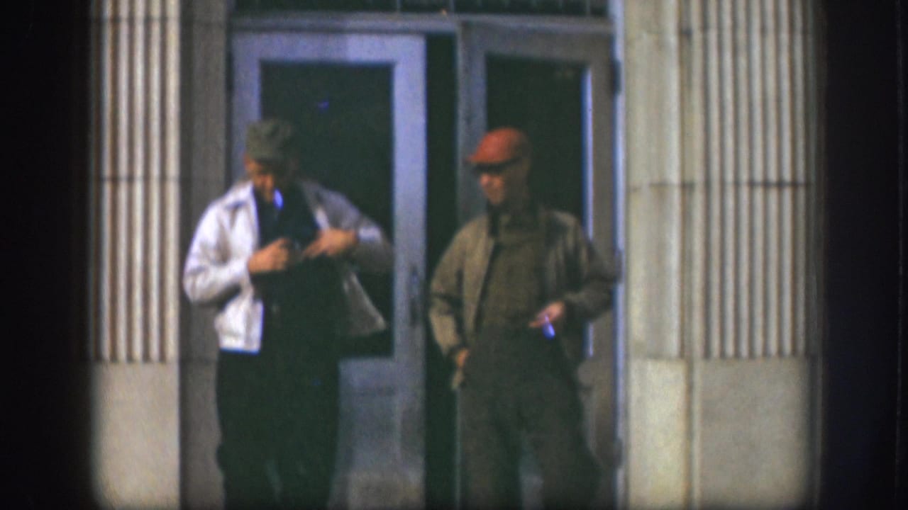Two men linger outside a building entrance in Kansas, circa 1958; the left man in a white jacket adjusts his clothing while the right in a red cap and olive jacket smokes.