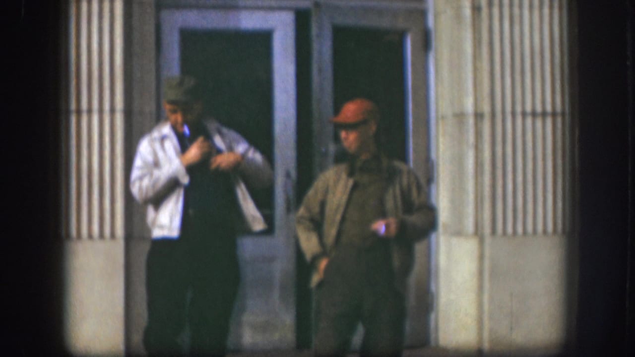 Two middle-aged men in caps outside a mid-century building in Kansas, circa 1958; one gestures while adjusting his white jacket, the other in a red cap and olive jacket holds a cigarette.