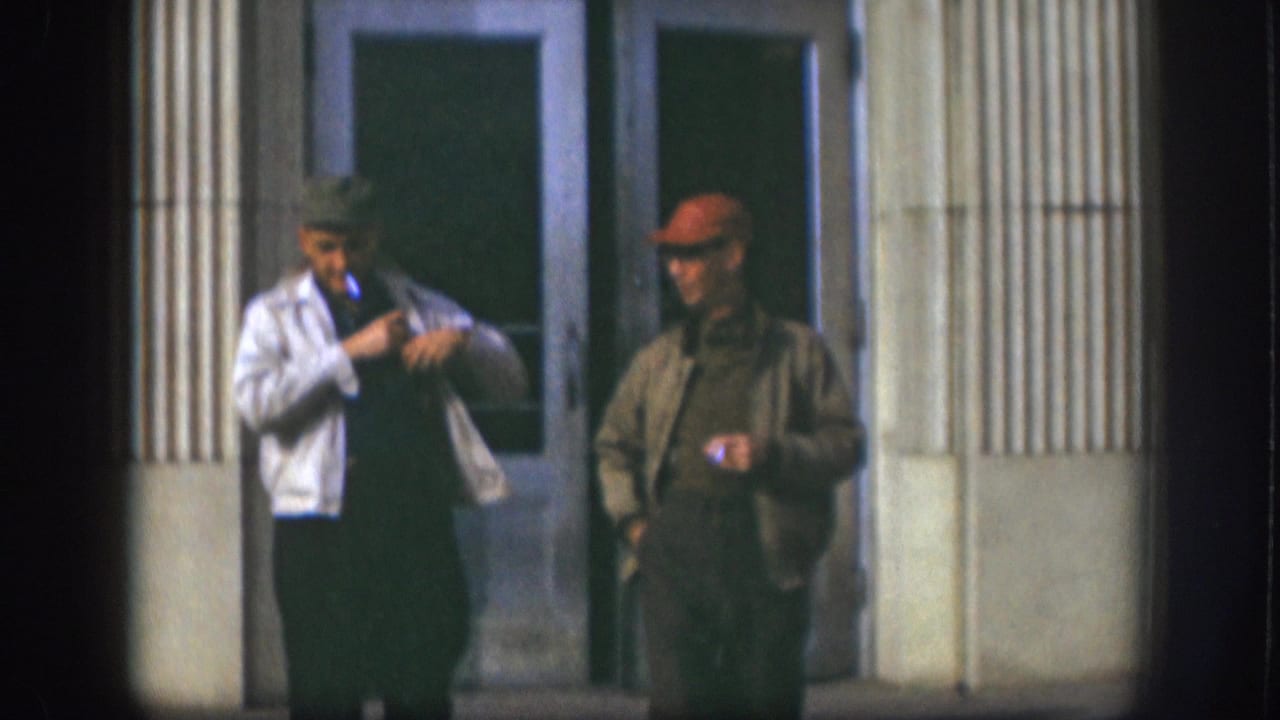 Two men in late-1950s workwear pause outside a fluted-column building facade in Kansas, USA, circa 1958; man in white jacket adjusts clothing while appearing to smoke.