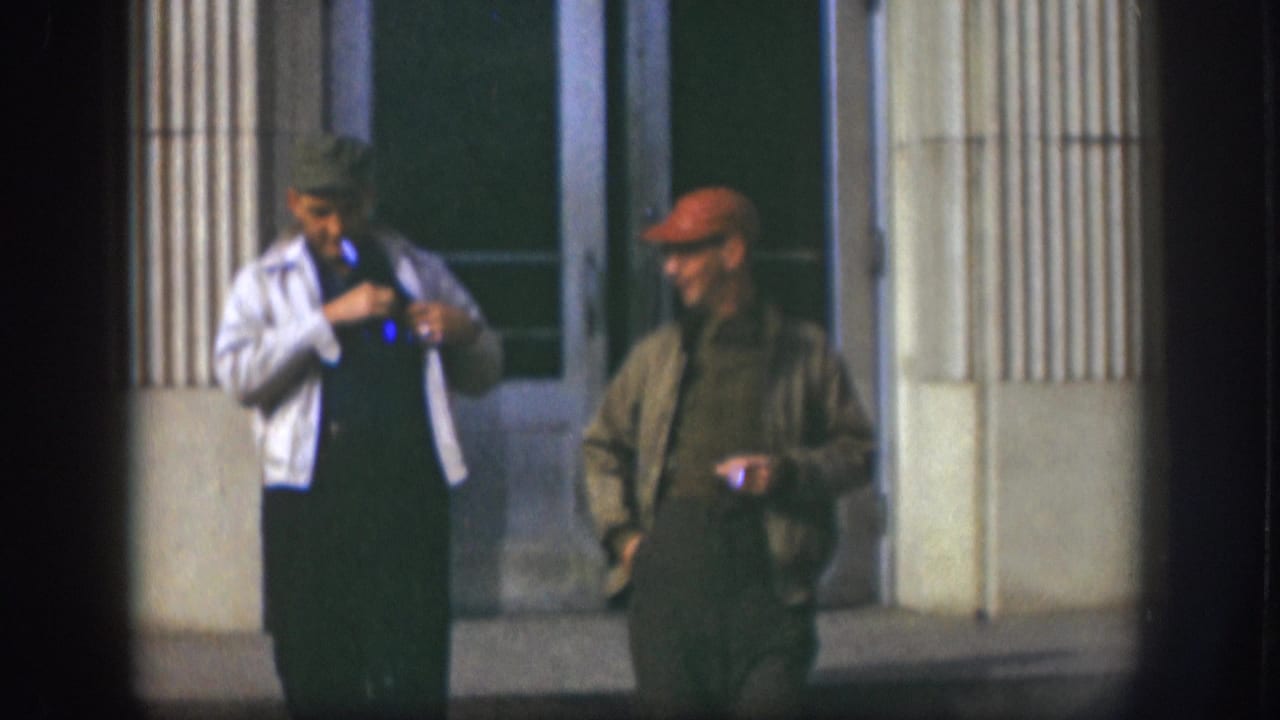 Two men outside a columned building; man on left gestures near his chest, man on right stands upright with hands at sides.