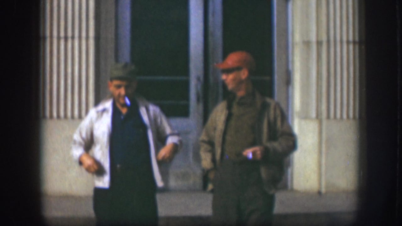 Two men in animated conversation outside a columned building in Kansas, USA, circa 1958; man in white jacket gestures expressively while companion in red cap faces him.
