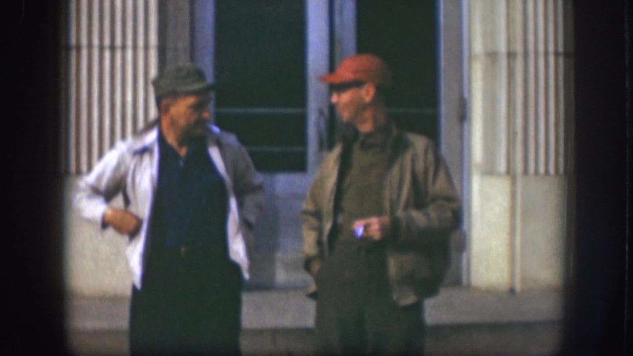 Two adult men in flat cap and red baseball cap conversing outside a columned building facade, Kansas USA, circa 1958.