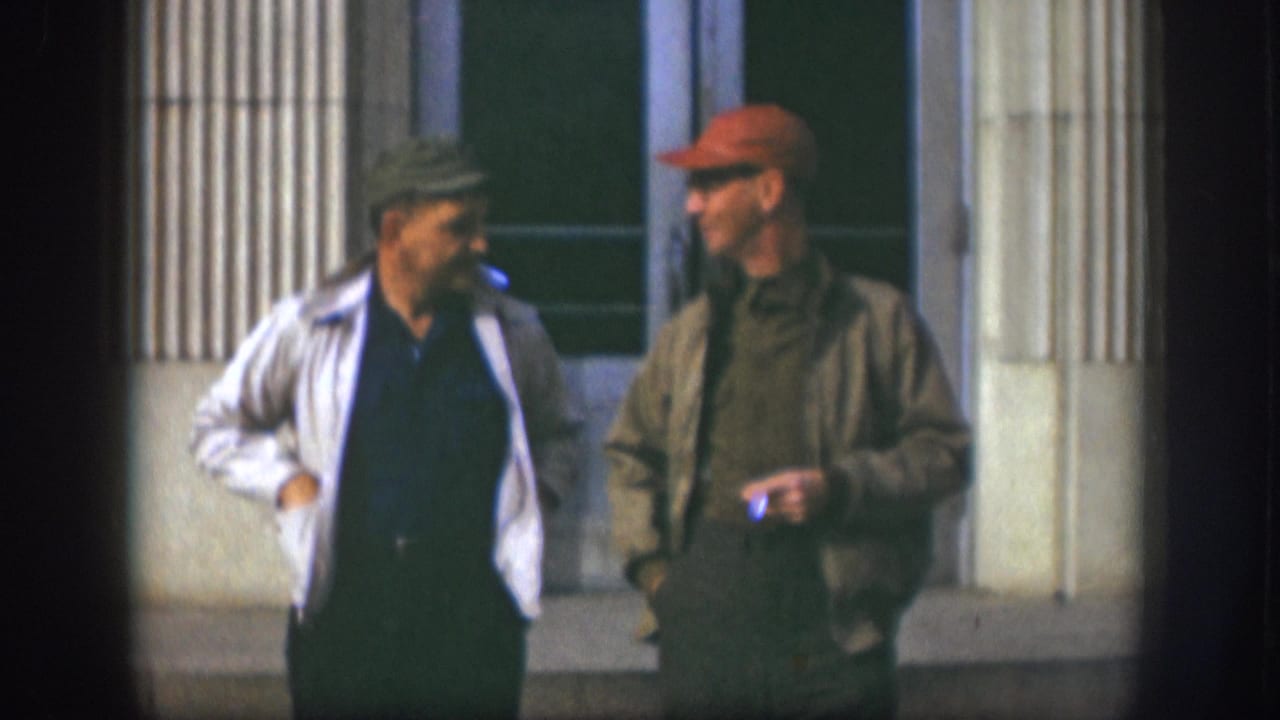 Two men mid-conversation outside a columned facade, one with arm raised in a casual gesture.