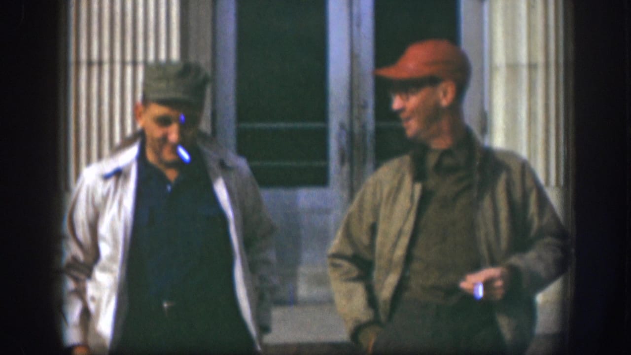 Two men in flat caps and casual jackets stand outside a substantial building entrance in what appears to be 1958 Kansas 8mm home movie footage.