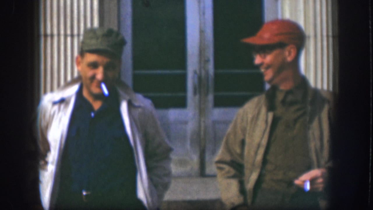 Two men in caps and jackets face each other outside a building with panelled double doors and stone columns; the left man has a cigarette.