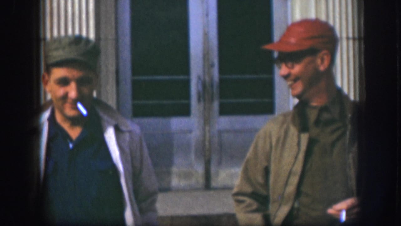 A candid close moment of two men laughing outside a building entrance; the man in a red cap and glasses is prominent in this 1950s 8mm home movie frame.