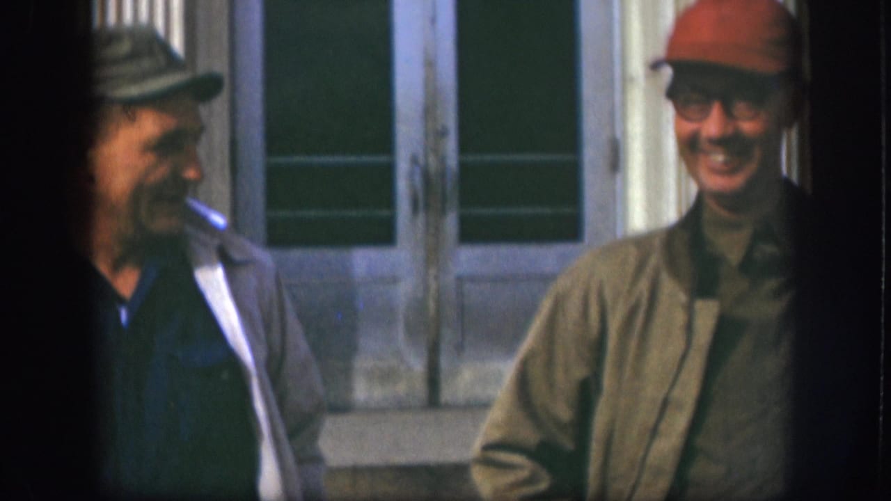 Two men share an open laugh outside a building entrance in this candid late-1950s 8mm home movie; one turns in profile with a cigarette while the other in a red cap smiles broadly.