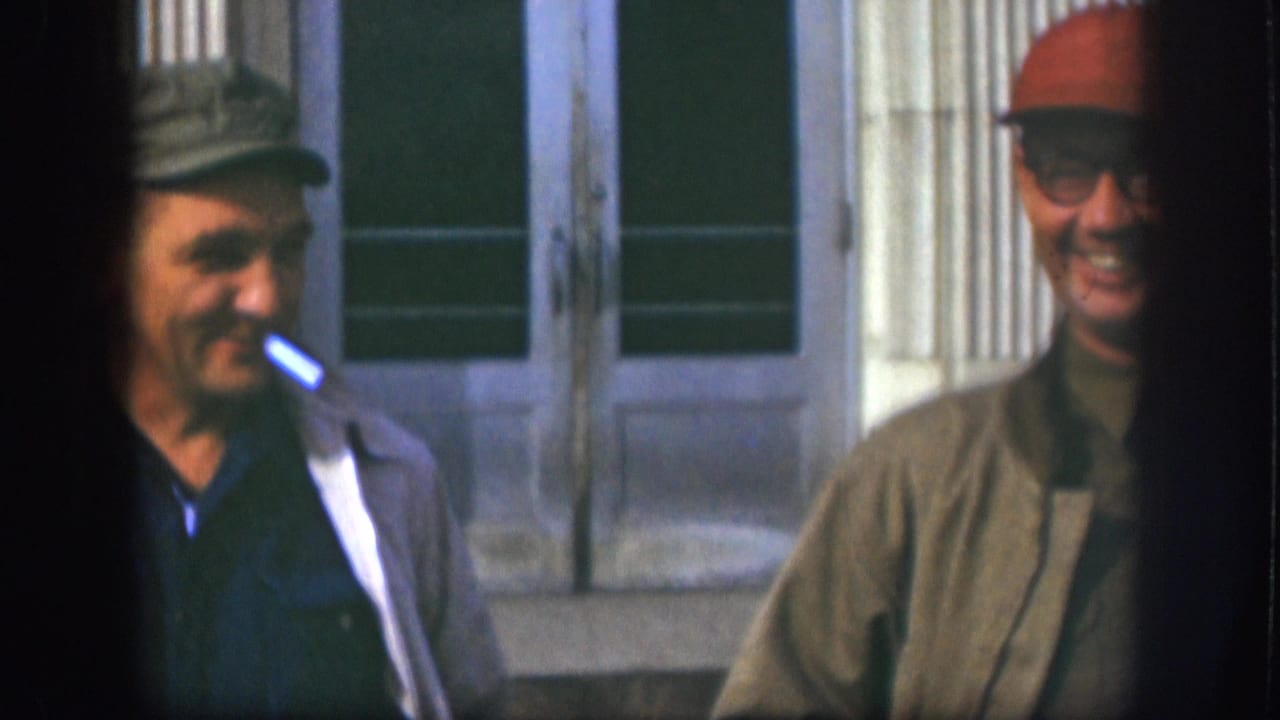 Close-up of two men in late-1950s casual jackets near a building; one smokes a cigarette while the other, wearing a red cap and glasses, grins directly at the camera.
