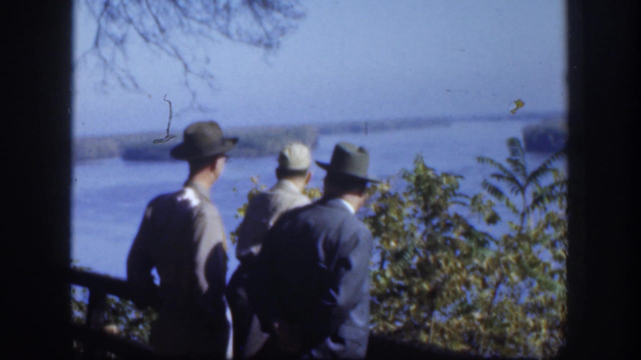 Several men in hats and jackets stand at what appears to be a fence or railing, viewed from behind, overlooking a broad pale lake with bare tree branches visible at frame left and shrubs in the foreground.