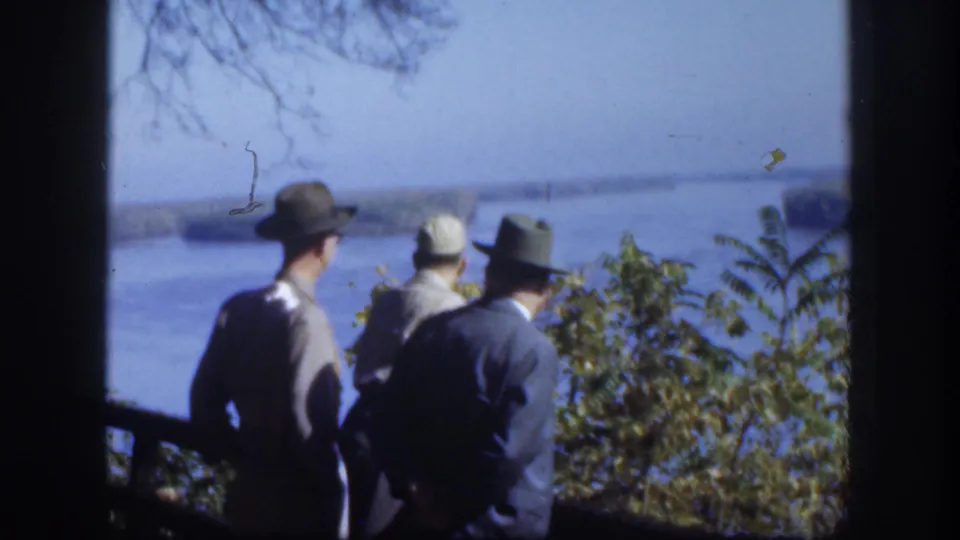 Several men in hats and jackets stand at what appears to be a fence or railing, viewed from behind, overlooking a broad pale lake with bare tree branches visible at frame left and shrubs in the foreground.