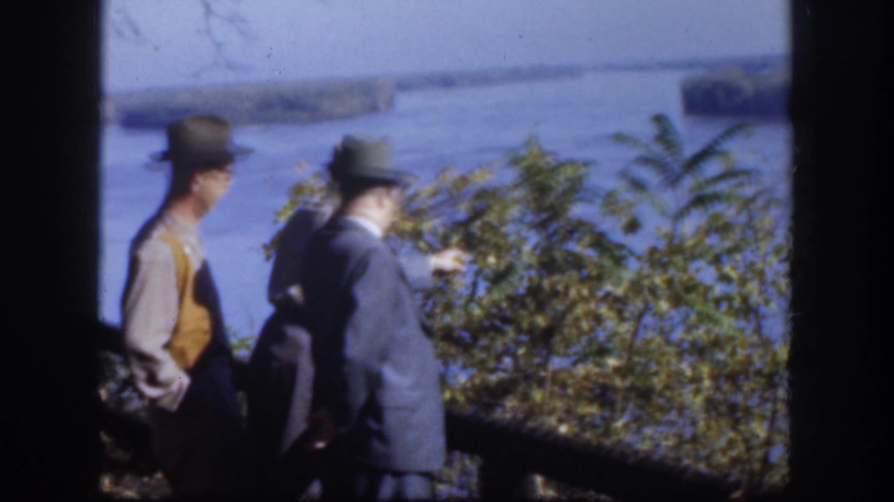 Two men — one in a yellow-ochre vest over a jacket, the other in a gray-blue jacket — stand near green shrubs looking out over a wide flat lake, consistent with the 1958 Kansas location metadata.