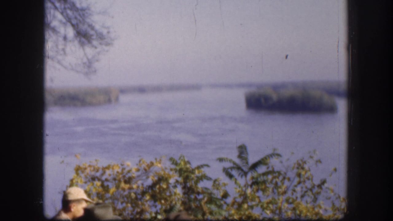 1958 8mm home movie frame showing a Kansas river with low-lying tree-covered islands; one man's hat visible at lower-left; yellow-green foreground shrubs.