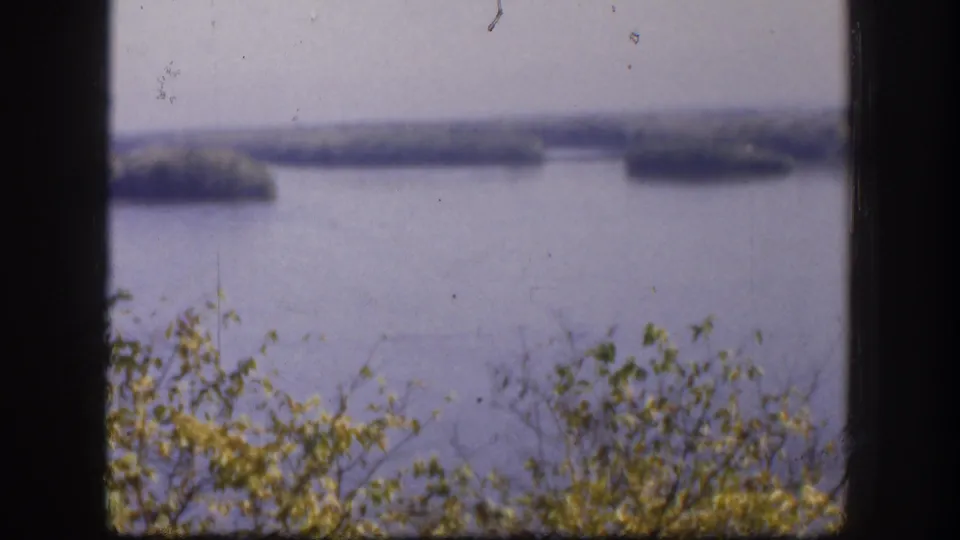 Final 8mm frame from 1958 Kansas home movie; river with scattered islands and foreground foliage; blue-lavender color shift from film aging.