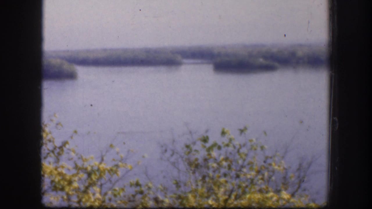 1958 8mm color home movie of a lake or reservoir in Kansas, USA, with distant wooded islands, shoreline shrubs, and a pale hazy sky.