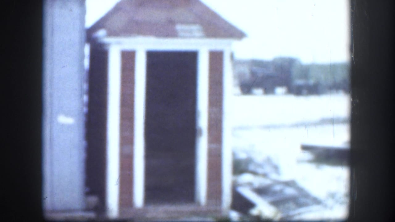 Frame from a 1958 Kansas 8mm home movie showing a small wooden outhouse with a reddish-brown pyramidal roof, white trim, and open dark doorway set in a snow-covered yard.
