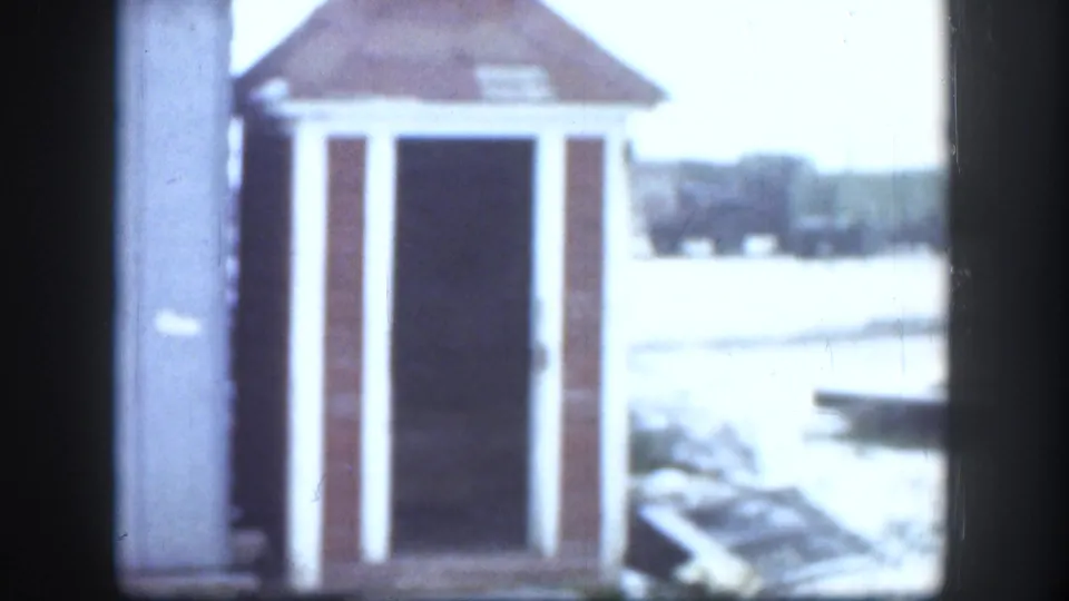 Frame from a 1958 Kansas 8mm home movie showing a small wooden outhouse with a reddish-brown pyramidal roof, white trim, and open dark doorway set in a snow-covered yard.