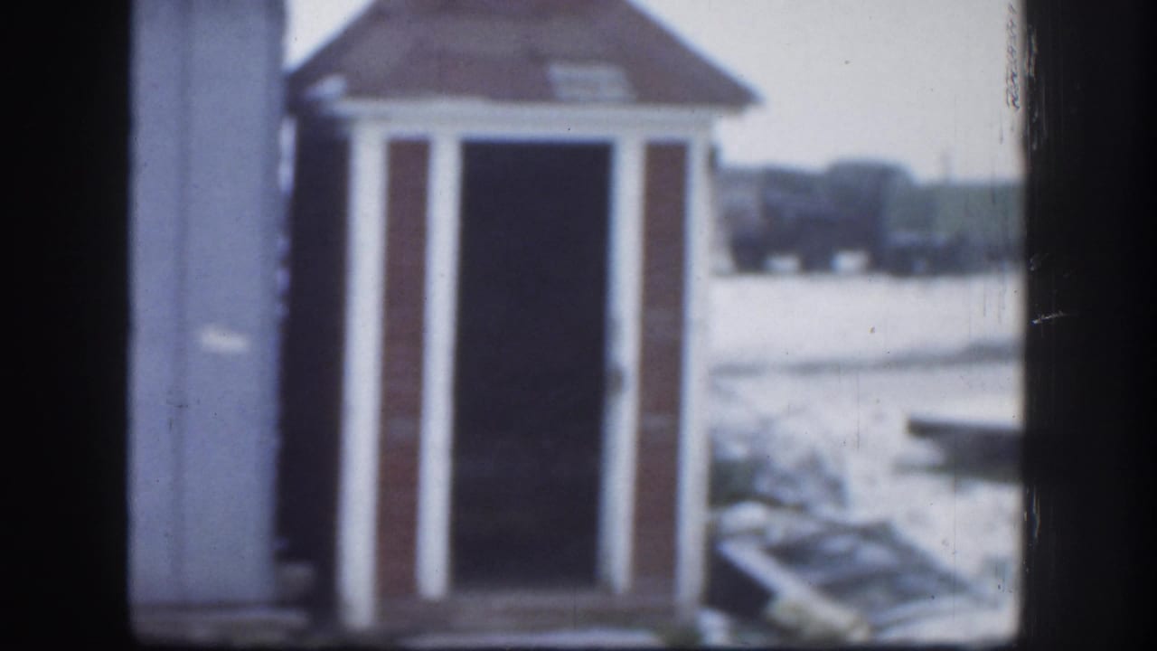 Second frame from 1958 Kansas 8mm clip showing outhouse with reddish-brown hip roof and white trim; pale overcast sky and faint background treeline visible.