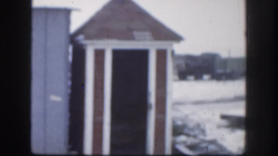 Mid-clip frame from 1958 Kansas home movie; wooden outhouse with pyramidal roof in snow, background shows dark shapes consistent with parked vehicles or farm equipment.
