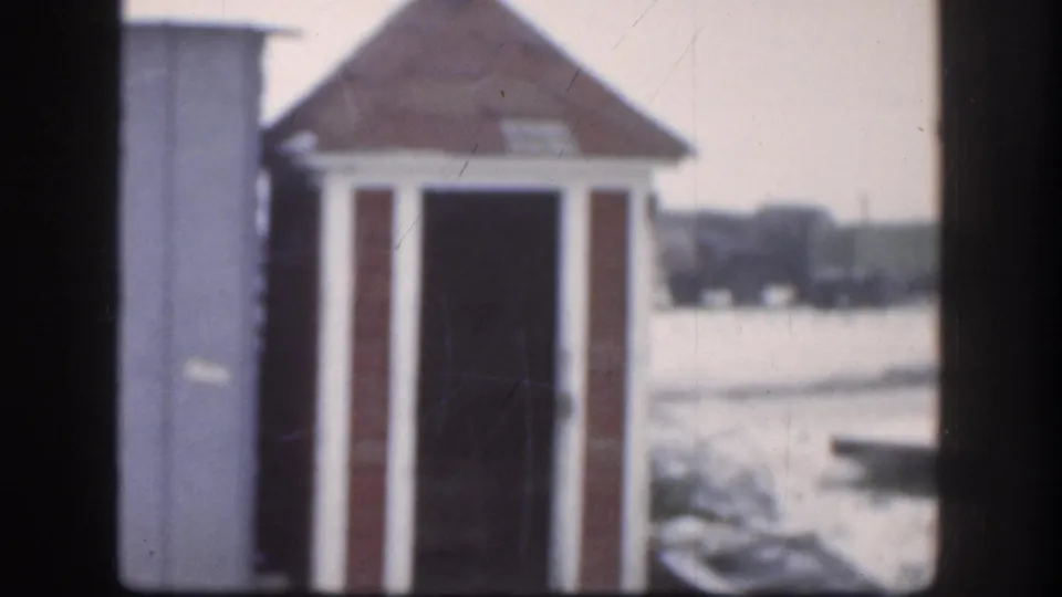 Closing frame of 1958 Kansas 8mm clip; wooden outhouse with pyramidal roof and white trim stands in snowy yard, faint background vehicles partially visible.
