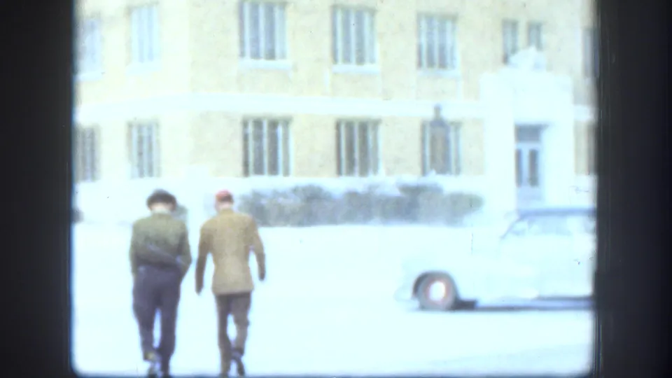 Two men walking away from camera across a snow-covered area near a multi-story institutional brick building and a parked car, Kansas USA circa 1958.