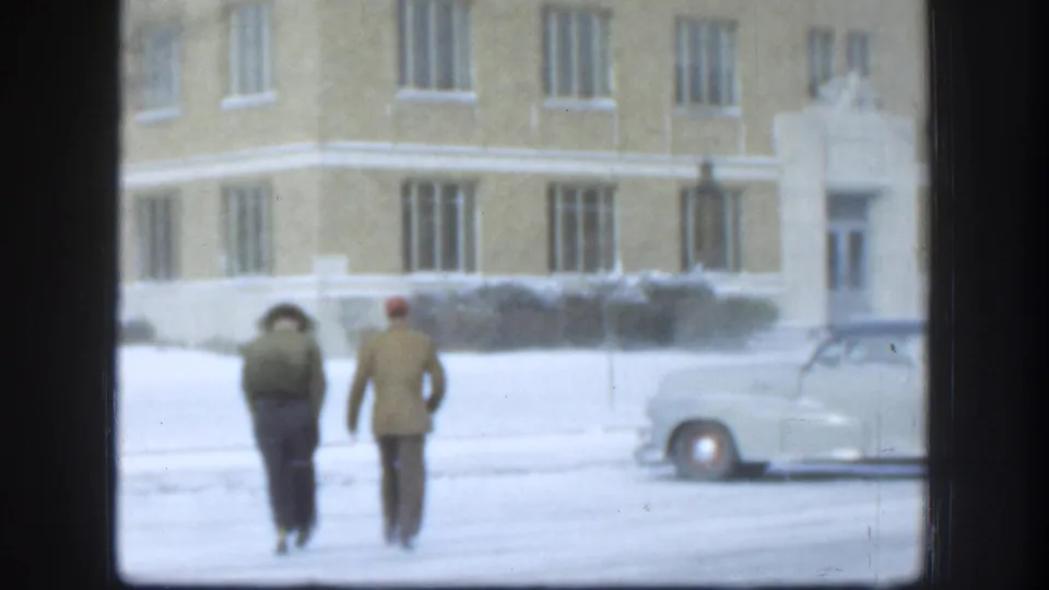 Two men in winter clothing walking across snowy ground past a parked sedan near the corner of a large tan brick building, Kansas USA circa 1958.