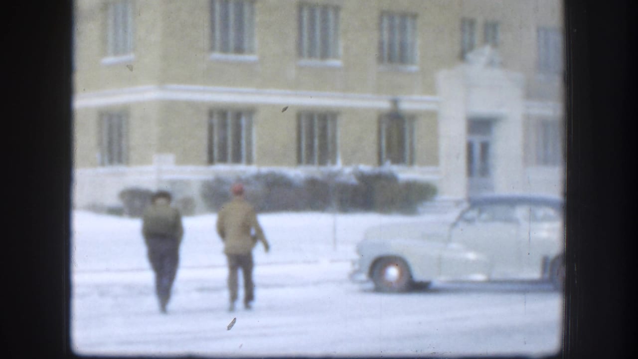 Two figures in period winter attire cross a snow-covered courtyard toward a large tan brick institutional building in Kansas, USA, circa 1958, framed by a dark interior window.