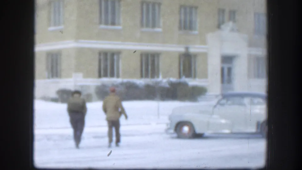 Two figures in period winter attire cross a snow-covered courtyard toward a large tan brick institutional building in Kansas, USA, circa 1958, framed by a dark interior window.