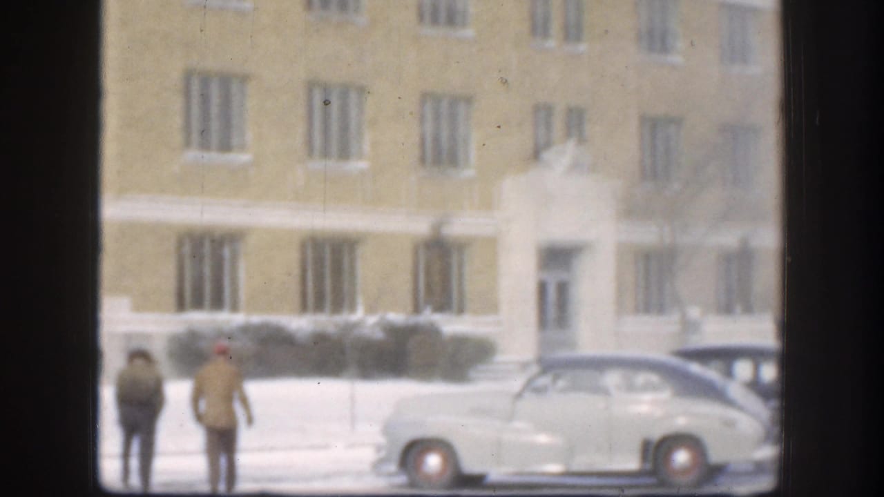 Two people standing near parked vintage sedans in front of a large tan brick institutional building in winter, seen through a window.