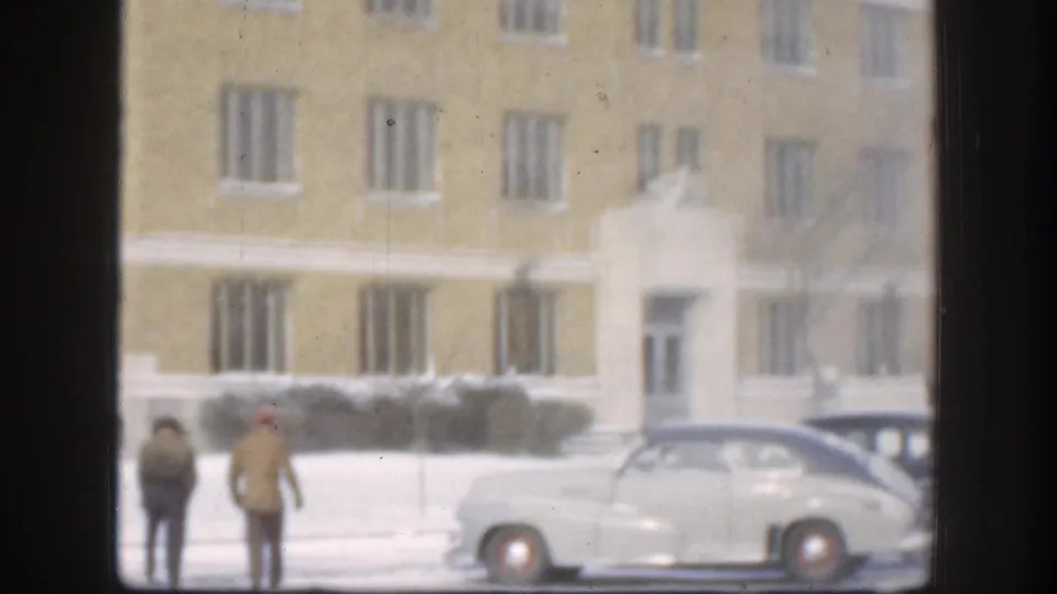 Two figures pause near two vintage sedans outside a large tan brick institutional building on a snowy day in Kansas, USA, circa 1958, framed by a dark window.