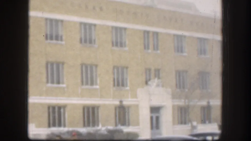 Multi-story tan brick institutional building with ornamental cornice detailing and a central entrance, snow at the base, Kansas, USA, circa 1958, viewed through a window frame.