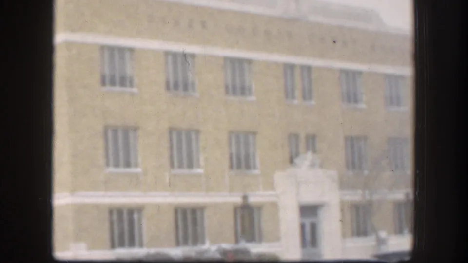 Large tan multi-story institutional brick building with small figures near the central entrance in winter, Kansas, USA, circa 1958, framed by an interior window.