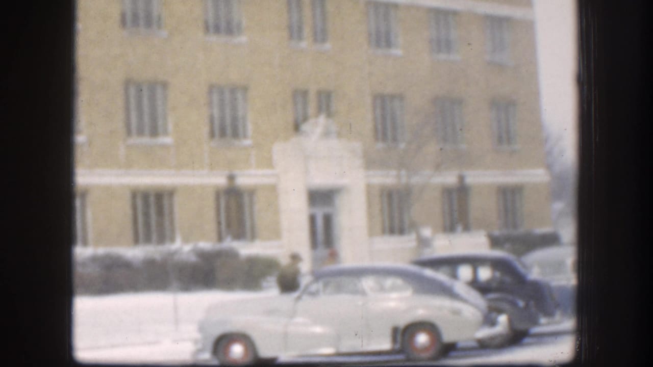 Circa 1958 Kansas winter: mid-century automobiles parked outside a large stone-and-brick institutional building, snow on the ground, a person visible at curbside.