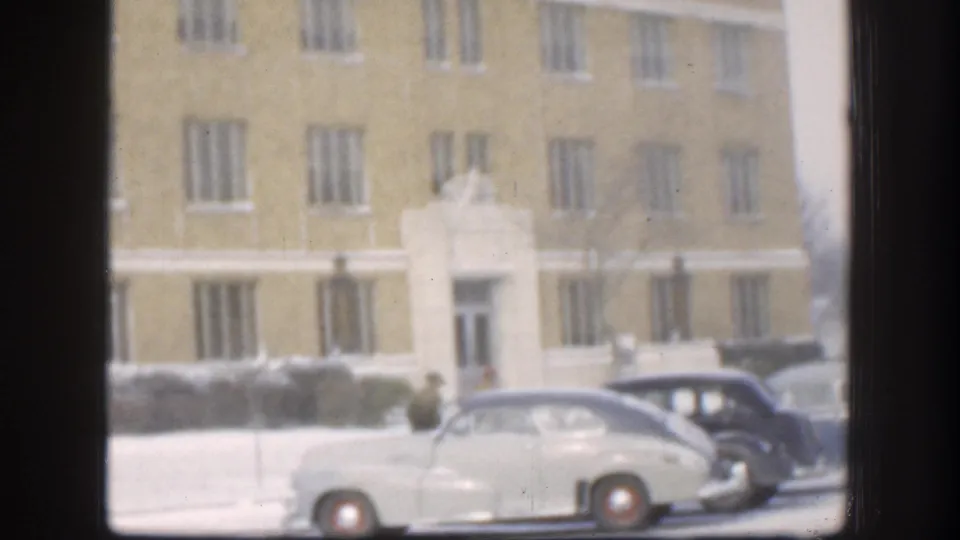 Circa 1958 Kansas winter: mid-century automobiles parked outside a large stone-and-brick institutional building, snow on the ground, a person visible at curbside.