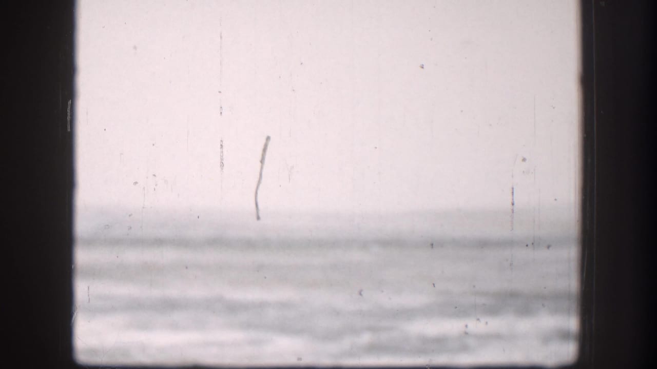 Continued 8mm view of flat plains through window, Kansas circa 1958; overcast sky and flat horizon with a faint vertical structure visible at right through washed-out exposure.