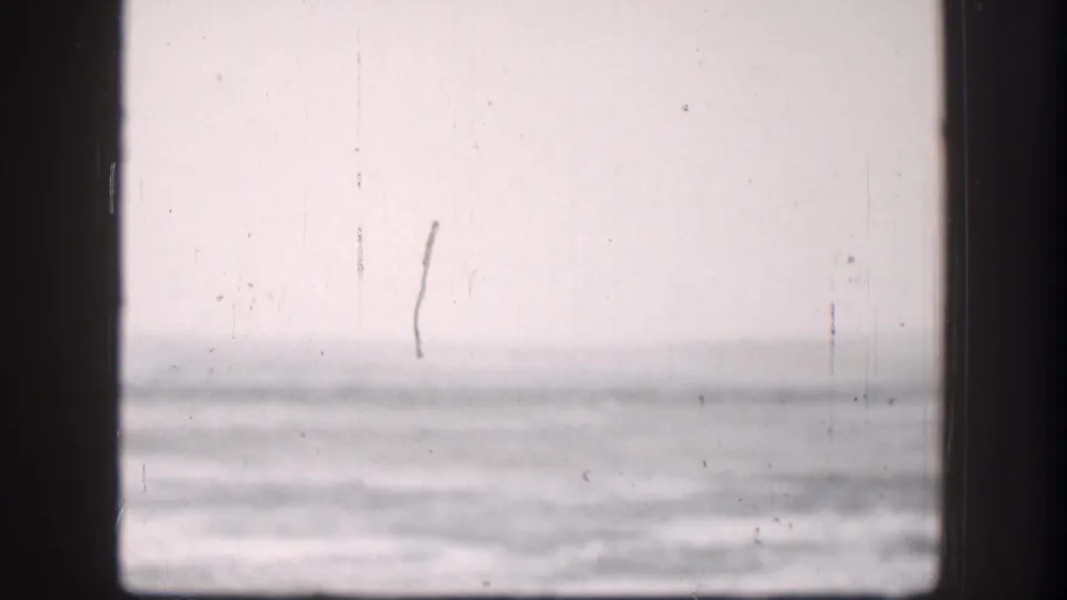 Continued 8mm view of flat plains through window, Kansas circa 1958; overcast sky and flat horizon with a faint vertical structure visible at right through washed-out exposure.