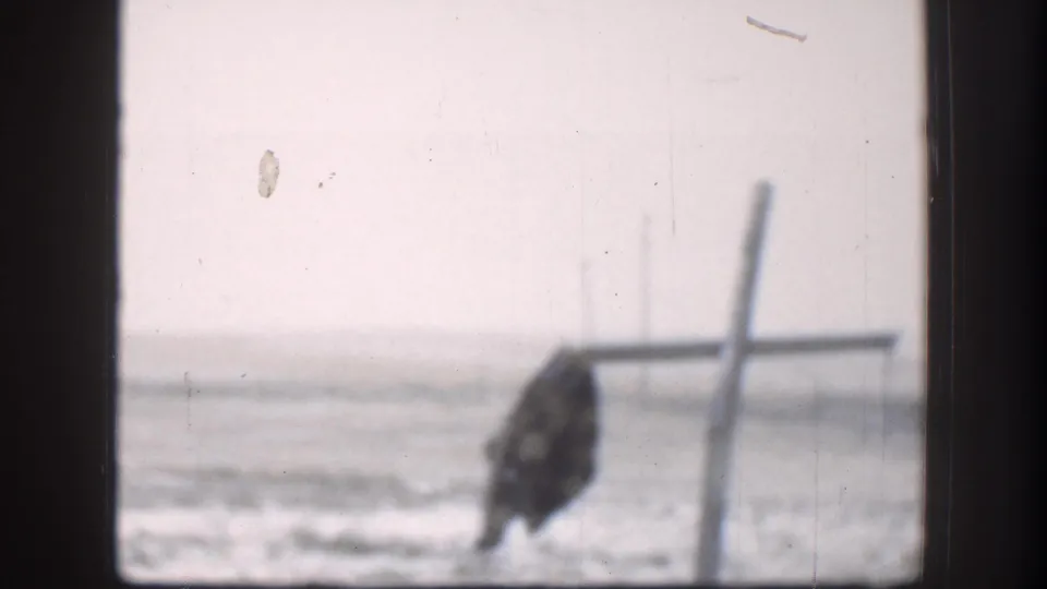 8mm shot through a window of Kansas plains circa 1958; a figure in dark clothing crouches near a wooden post or fence in an open field under an overcast sky.