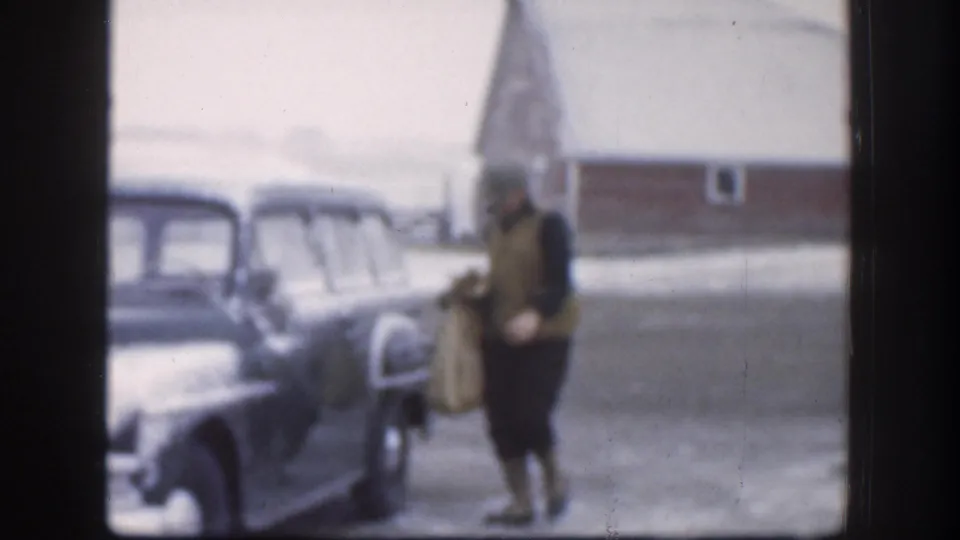Two adults beside a dark mid-1950s American sedan near a red outbuilding, Kansas circa 1958; shot through a window with film grain and muted tones throughout.