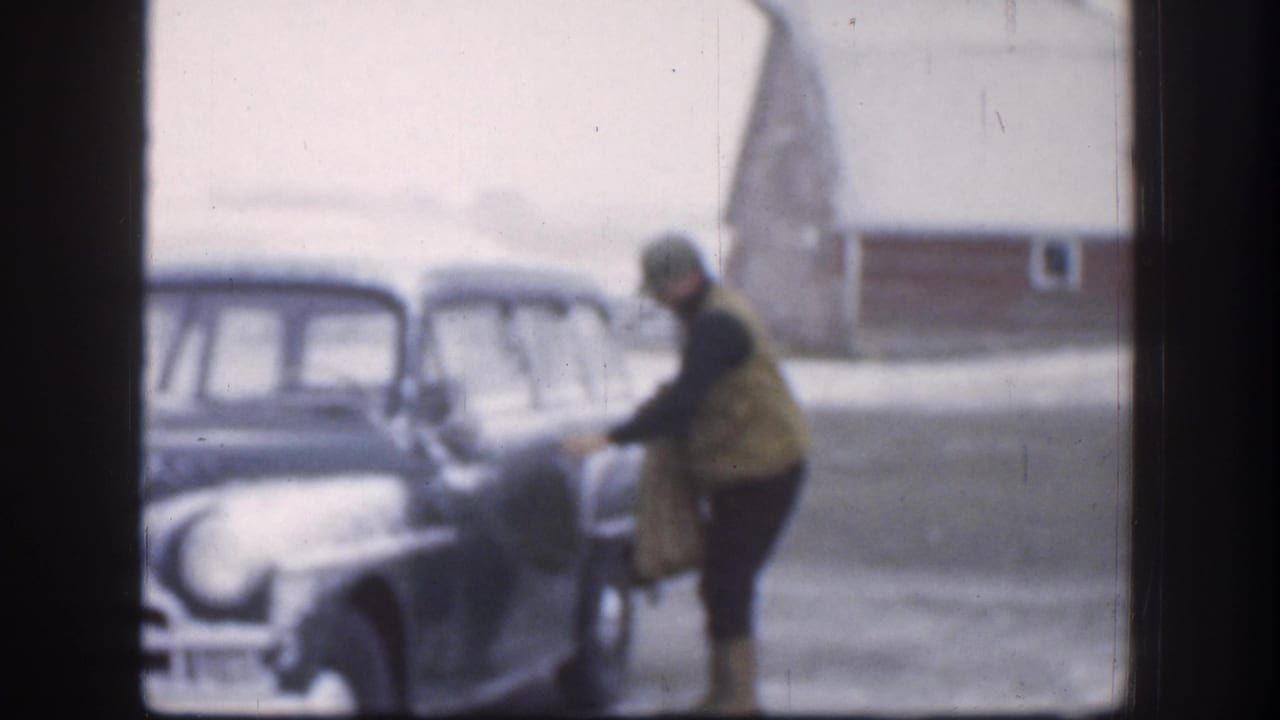 Adult in tan winter jacket approaching a large dark American sedan in a snowy Kansas farmyard, red barn visible behind, circa 1958 8mm home movie.