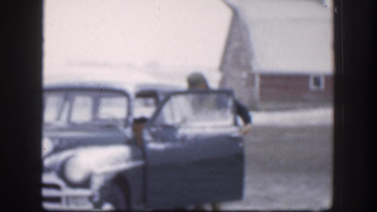 Two adults at the open door of a large 1950s American sedan in a snowy rural Kansas farmyard, red barn behind, circa 1958 8mm footage.