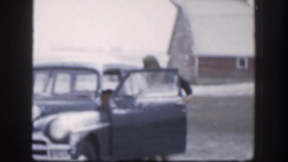 Two adults at the open door of a large 1950s American sedan in a snowy rural Kansas farmyard, red barn behind, circa 1958 8mm footage.