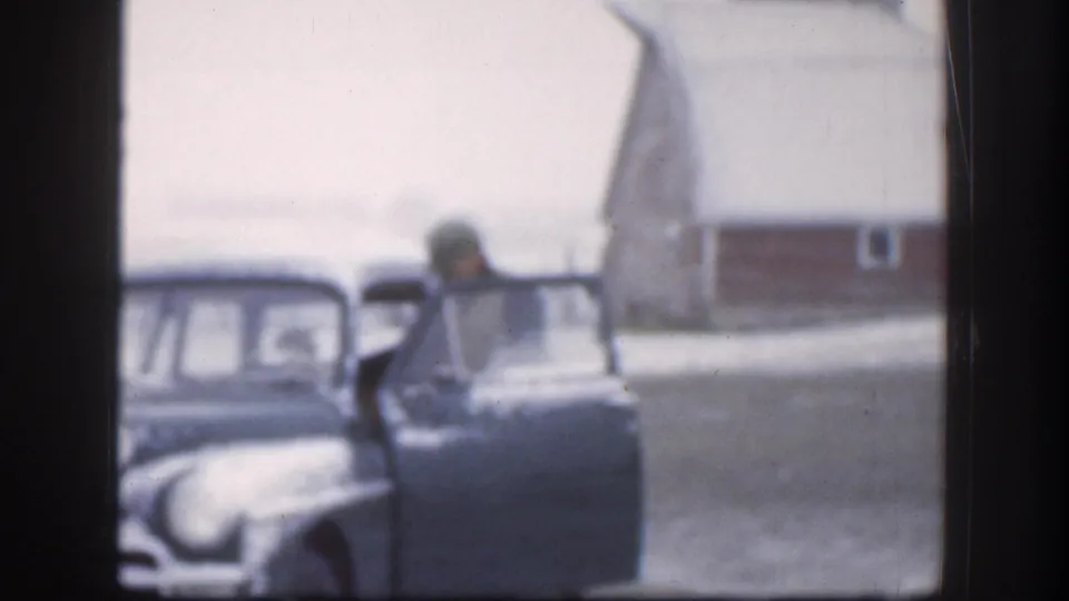 Person in winter hat leaning toward open door of a 1950s American sedan on a snow-dusted Kansas farmstead, circa 1958, 8mm film.